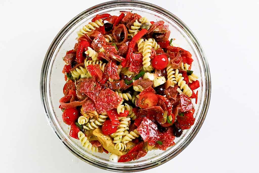 A glass bowl filled with Pasta Salad inspired by Italian Antipasto—rotini noodles, sliced salami, cherry tomatoes, black olives, mozzarella balls, red peppers, and herbs—makes for an easy recipe set against a white background.