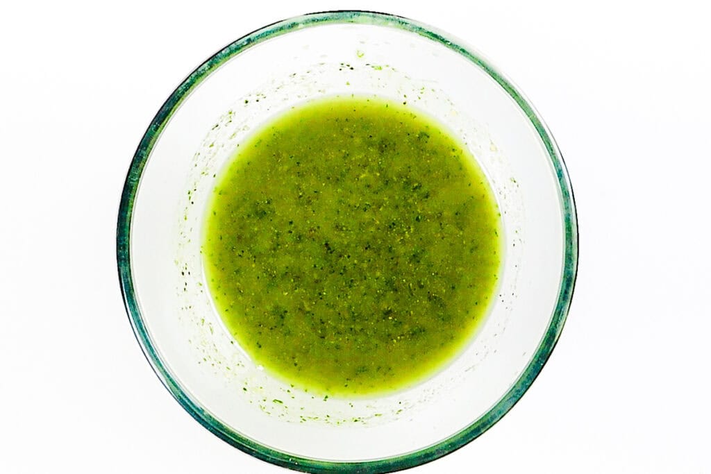 A clear glass bowl filled with green salsa, viewed from above, against a white background. The salsa appears smooth with a slightly chunky texture and flecks of herbs, making it an easy recipe to pair with chips or as a zesty pasta salad topping.