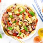 A bowl of Italian Antipasto pasta salad with cherry tomatoes, olives, mozzarella, red onions, basil, artichokes, and cured meat. Fresh basil, cherry tomatoes, and oil bottles surround this easy recipe for a vibrant meal.