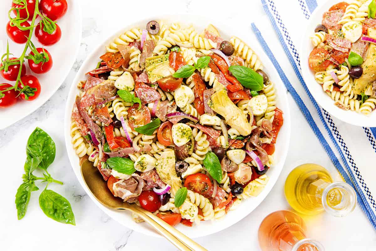 A bowl of Italian Antipasto pasta salad with cherry tomatoes, olives, mozzarella, red onions, basil, artichokes, and cured meat. Fresh basil, cherry tomatoes, and oil bottles surround this easy recipe for a vibrant meal.