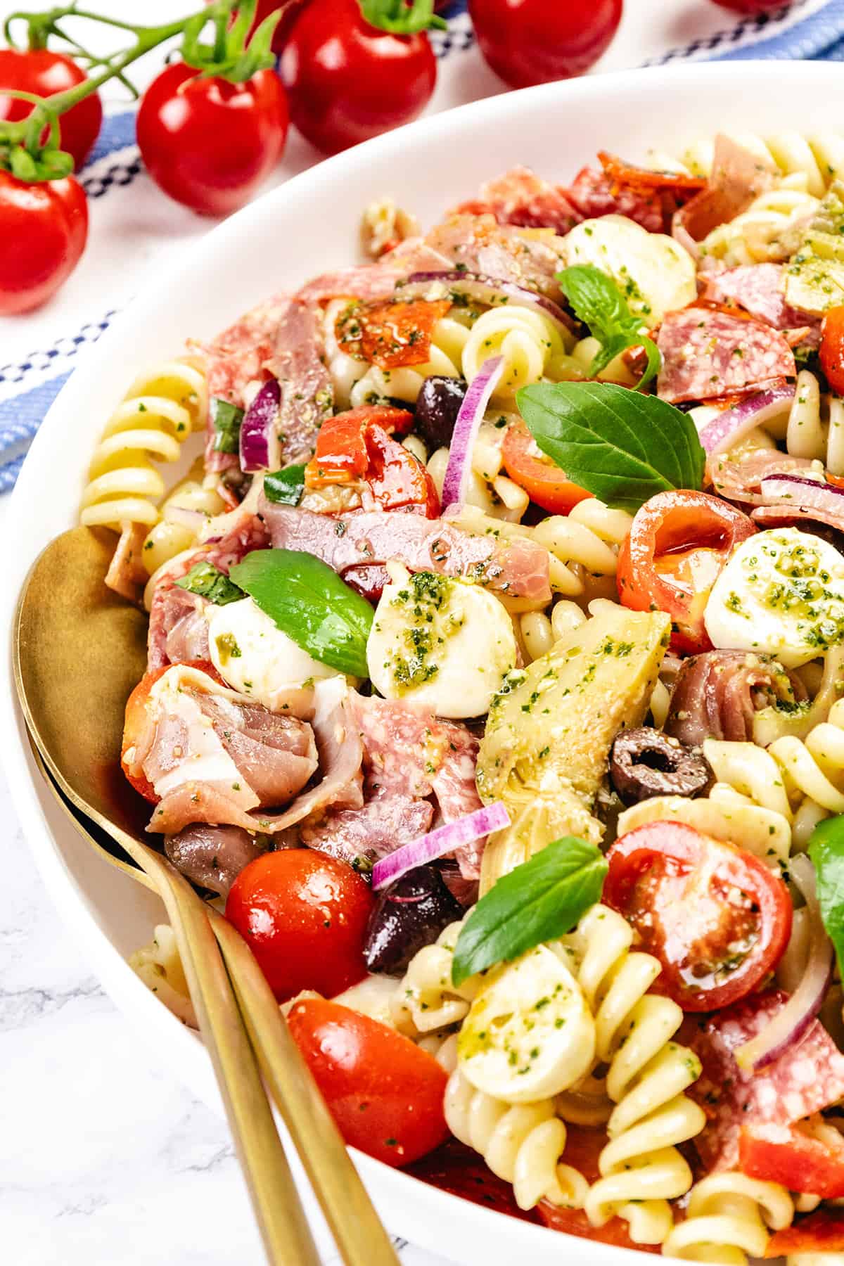 A bowl of Italian Antipasto pasta salad with cherry tomatoes, mozzarella balls, red onion, olives, salami, basil leaves, and artichoke hearts, garnished with pesto. A gold fork and spoon are placed in the bowl for this easy recipe.