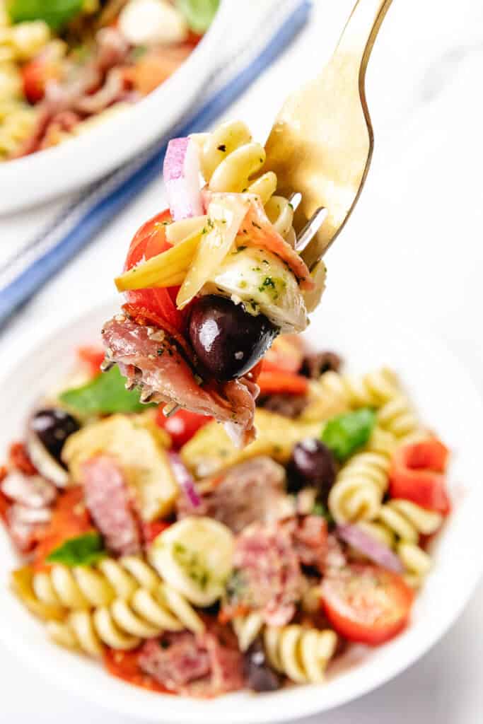 A fork holds a bite of Italian Antipasto pasta salad with rotini, black olive, red onion, tomato, mozzarella, and cured meat above a bowl filled with the same colorful pasta salad—an easy recipe for any occasion.