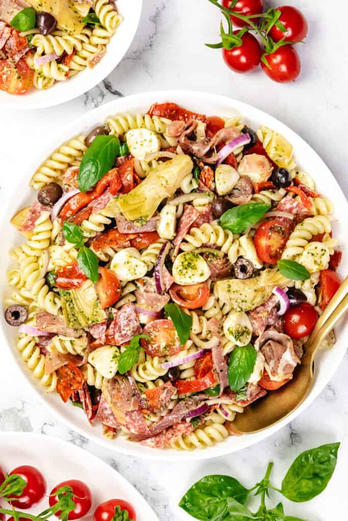 A white bowl filled with Italian Antipasto pasta salad, featuring rotini, cherry tomatoes, black olives, mozzarella balls, artichoke hearts, red onion, salami slices, basil leaves, and a green herb dressing. Fresh tomatoes and basil sit nearby.