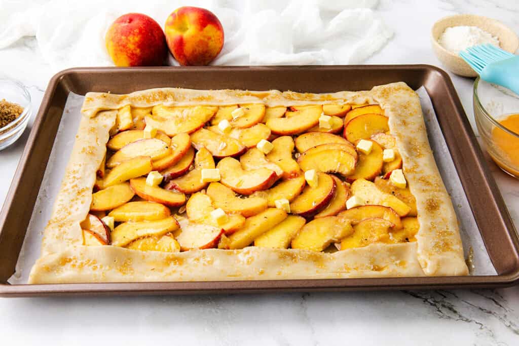 A rectangular Peach Galette made with flaky pie dough is topped with sliced peaches and butter cubes on a baking sheet. Two whole peaches and baking utensils rest in the background on a white counter.