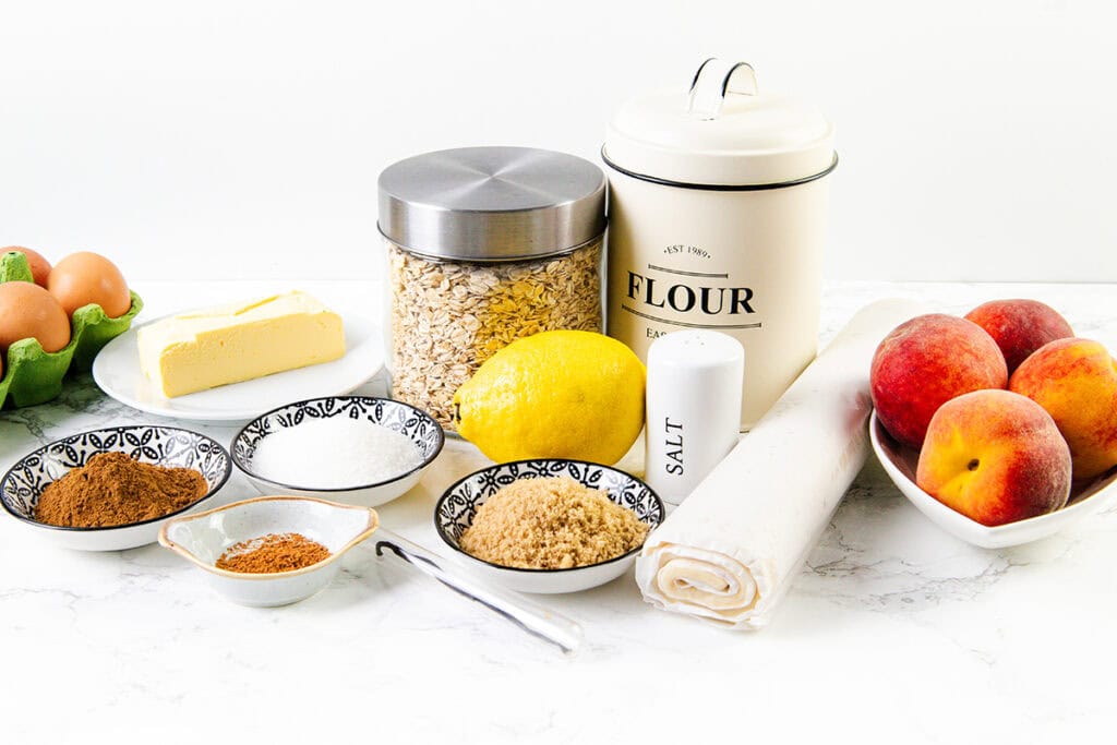 A baking setup with eggs, butter, oats, flour, peaches, brown sugar, cinnamon, nutmeg, lemon, salt, and a rolled Pie Dough sheet on a white surface. Ingredients are in bowls or containers.