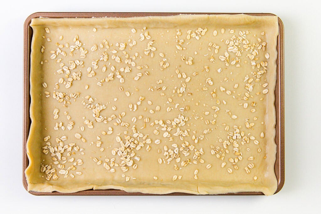 Rectangular baking tray lined with rolled-out pie dough, edges folded up, sprinkled with uncooked oats on top, viewed from above on a white background.