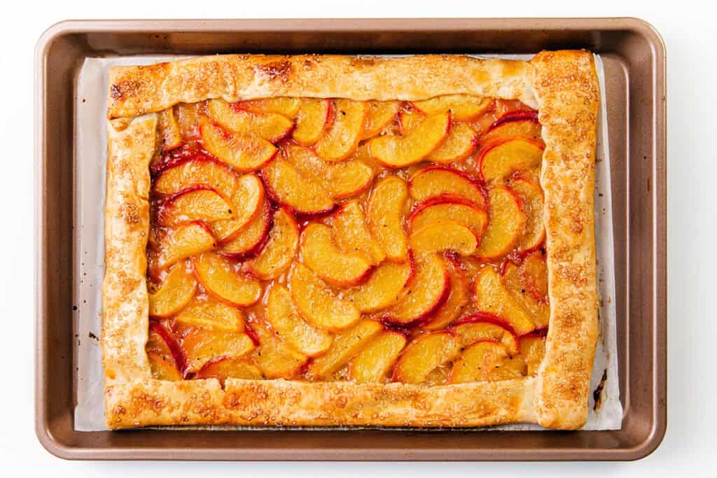 A rectangular peach galette made with golden, flaky pie dough and neatly arranged peach slices sits on a parchment-lined baking sheet.