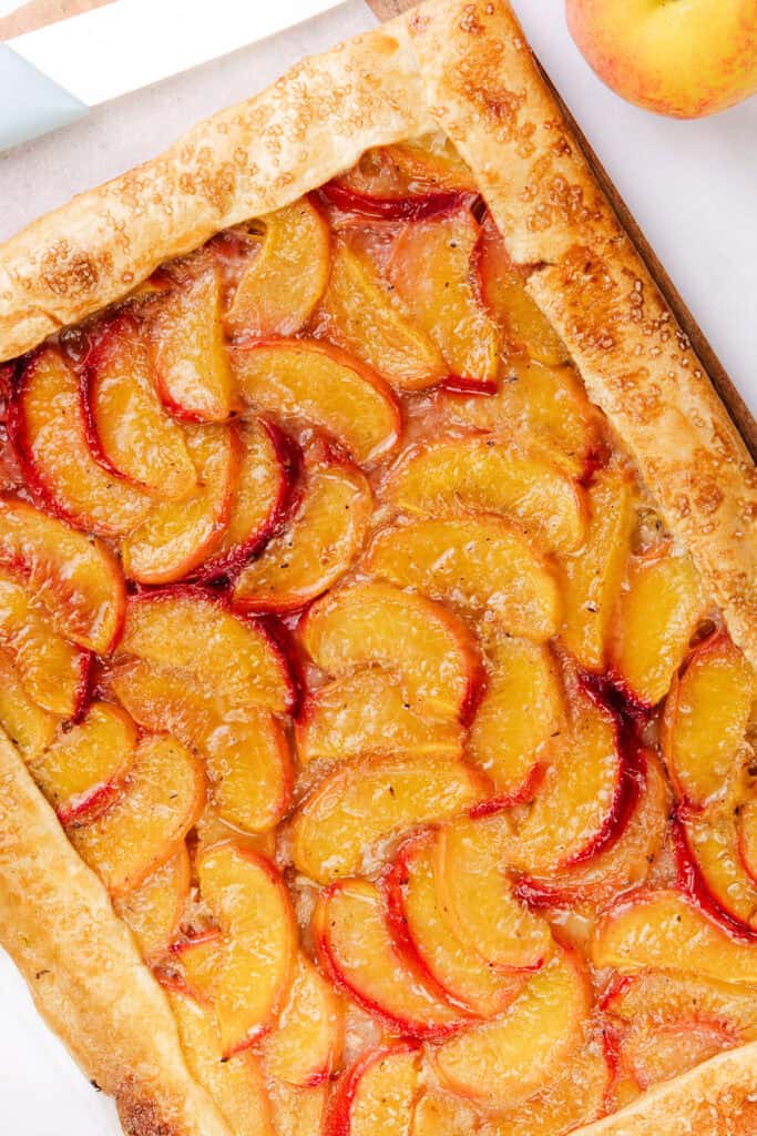 A rectangular peach galette with a golden, flaky pie dough crust, topped with neatly arranged slices of fresh peaches, sits on parchment paper next to a whole peach.