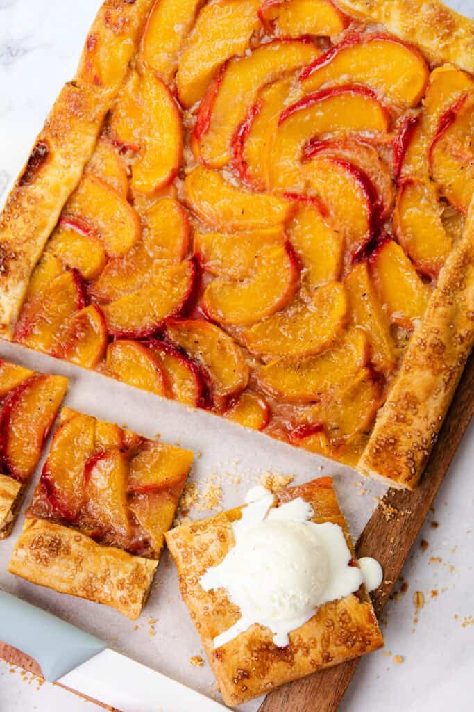 A rectangular Peach Galette with golden pie dough crust is topped with sliced peaches and sits on parchment paper. Two square pieces are cut, one finished with a scoop of melting vanilla ice cream.