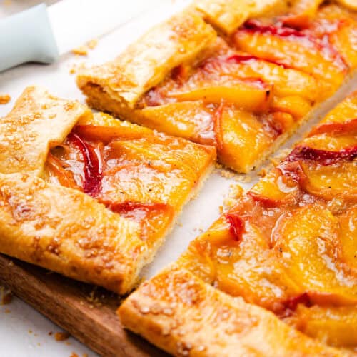 A close-up of a rectangular peach galette with golden, flaky puff pastry crust and glossy peach slices, partially cut into squares, rests on a wooden board with a knife nearby.