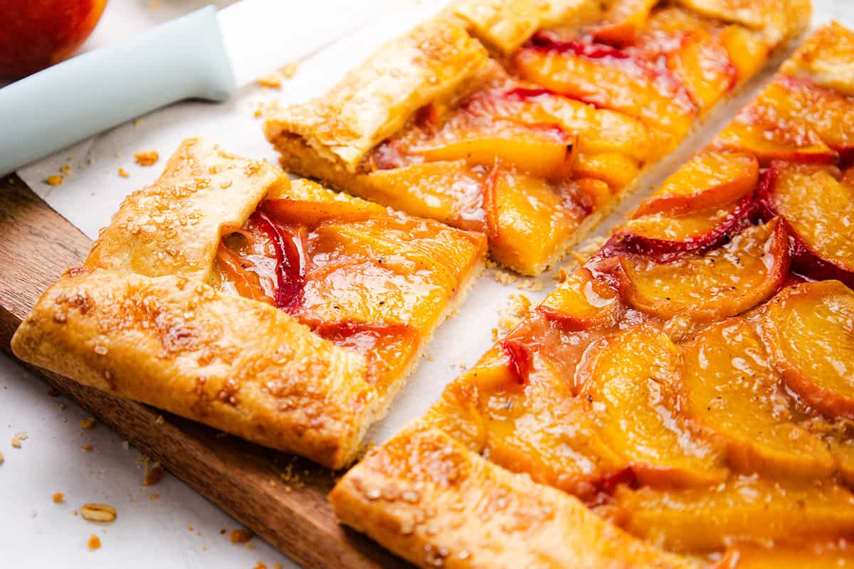 A close-up of a rectangular peach galette with golden, flaky puff pastry crust and glossy peach slices, partially cut into squares, rests on a wooden board with a knife nearby.