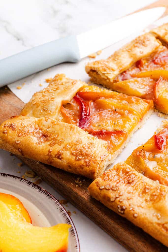 A close-up of a rustic Peach Galette with golden crust and peach slices, cut into squares on a wooden board. Made with flaky pie dough, it sits beside a knife and a plate holding an extra slice.
