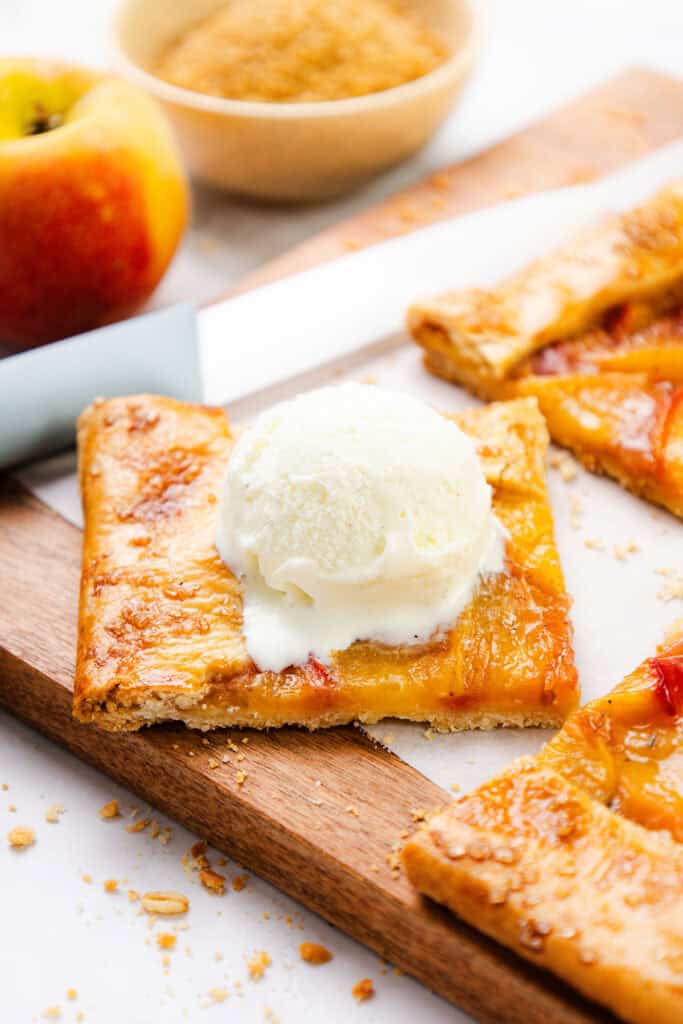 A square slice of apple tart made with flaky pie dough is topped with vanilla ice cream on a wooden board. Nearby, more tart slices, a fresh apple, a knife, and a bowl of brown sugar complete the scene.