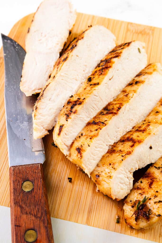 Sliced grilled chicken breast on a wooden cutting board with a large kitchen knife beside it. The juicy, brined chicken breast is perfectly browned with bold grill marks.