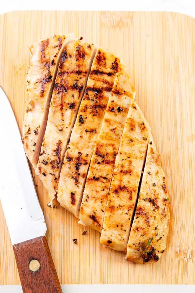 A grilled chicken breast sliced into strips on a wooden cutting board, with a knife resting beside it. The brined chicken breast shows perfect grill marks and is seasoned with herbs.