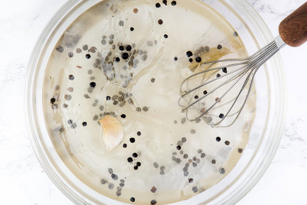 A glass bowl filled with clear liquid, whole black peppercorns, a garlic clove, and a whisk with a wooden handle, set on a white surface—ideal for preparing brined chicken breast for the perfect grilled chicken.