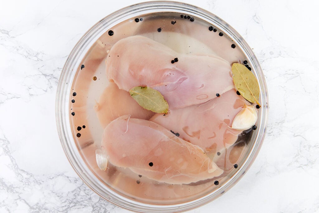 Raw chicken breasts soaking in a glass bowl of water with black peppercorns, bay leaves, and a garlic clove, are ready to become brined chicken—the secret to perfect grilled chicken every time. Set on a white marble surface.