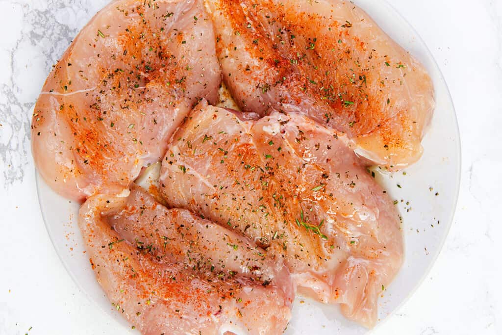 Raw brined chicken breast on a white plate, seasoned with paprika, black pepper, and green dried herbs, ready for the grill, sits on a marble surface.
