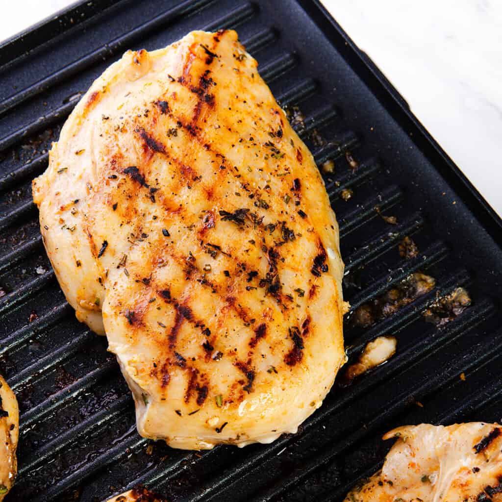 A perfect grilled chicken breast with visible grill marks and seasoning sits on a black grill pan, showcasing a cooked, golden-brown surface.