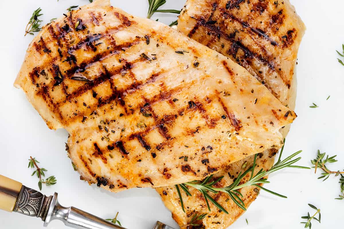 Grilled chicken breast with visible char marks, garnished with fresh rosemary and thyme, served on a white plate with a fork—perfect grilled chicken for any occasion.