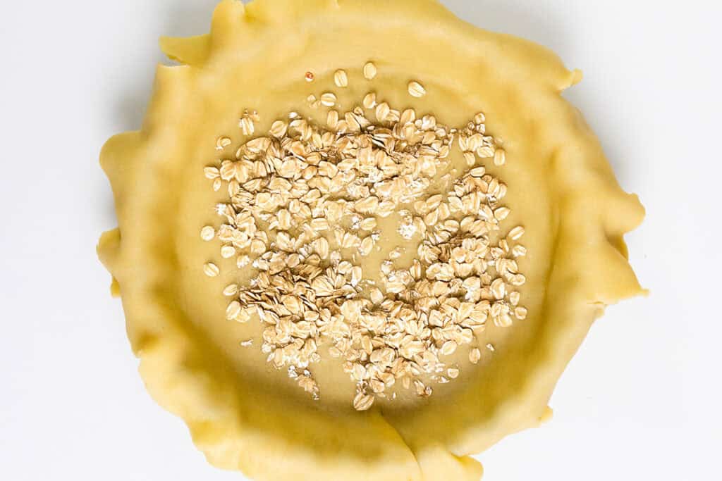 A flaky butter crust with crimped edges is placed on a white surface and filled with a layer of uncooked rolled oats.