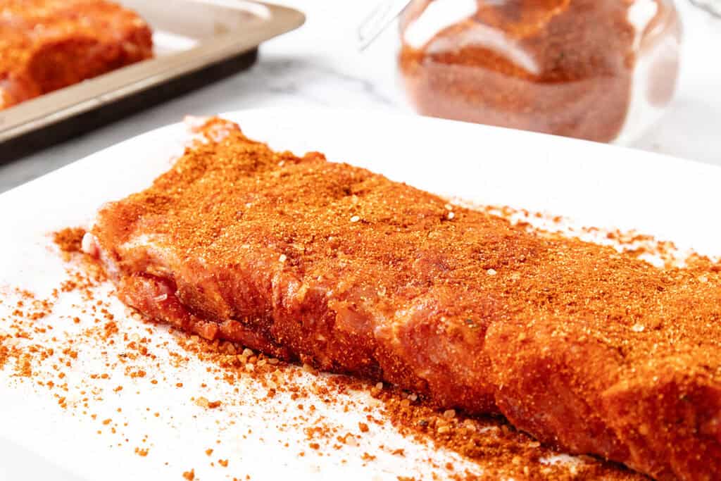 A rack of raw pork ribs on a white plate, coated with a reddish-brown dry rub seasoning, with extra seasoning scattered around and a jar of rub in the background.