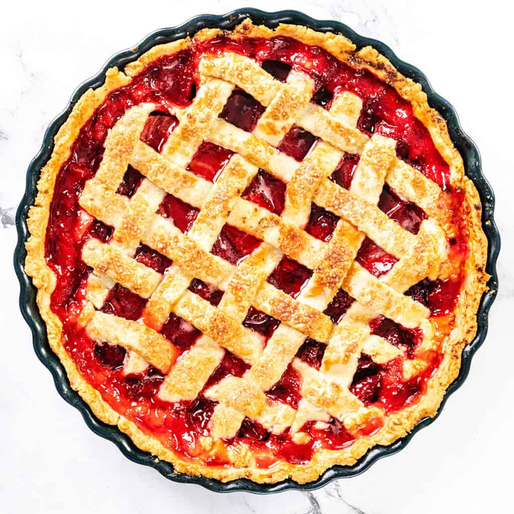 A baked Strawberry Pie with a golden-brown flaky butter crust sits in a pie dish. The red strawberry filling peeks through the woven pastry strips, and sugar crystals are sprinkled on top.