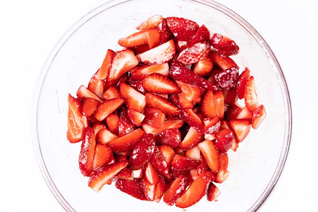 A clear glass bowl filled with sliced fresh strawberries, coated with sugar, perfect for preparing a classic strawberry pie or topping a flaky butter crust, on a white background.