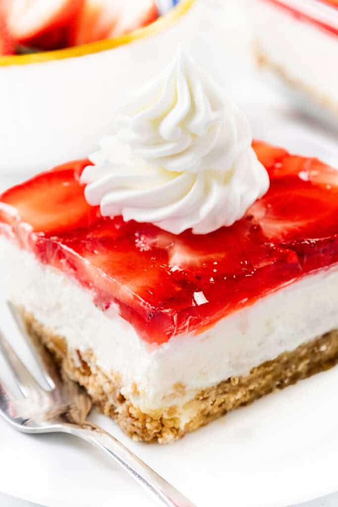 A slice of strawberry cheesecake with a graham cracker crust, creamy filling, strawberry topping reminiscent of strawberry pretzel salad, and a swirl of whipped cream on top, served on a white plate with a fork.