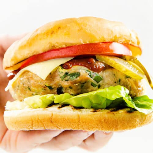 A hand holding a sandwich with lettuce, a moist turkey burger patty, cheese, tomato slices, pickles, and ketchup inside a toasted burger bun, against a white background.