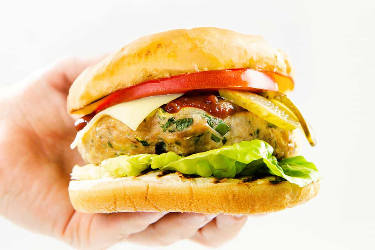A hand holding a sandwich with lettuce, a moist turkey burger patty, cheese, tomato slices, pickles, and ketchup inside a toasted burger bun, against a white background.