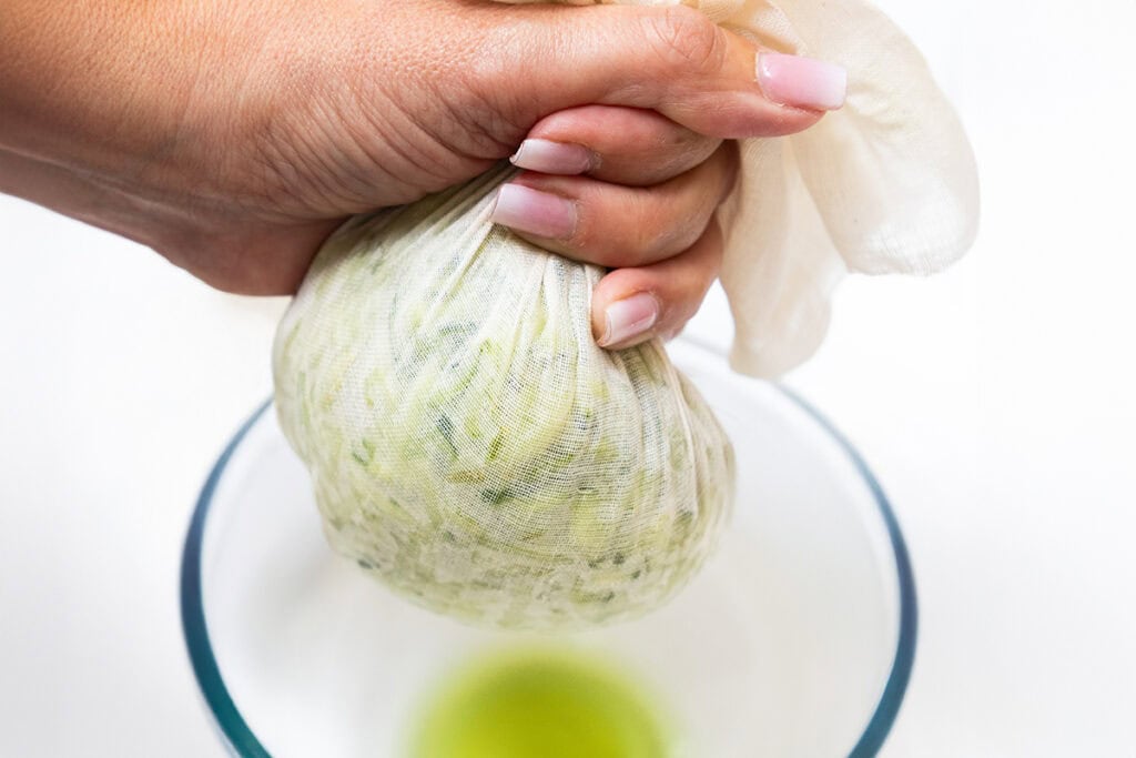 A hand squeezes shredded zucchini in a cheesecloth over a glass bowl, extracting green liquid—an essential step for making super moist burgers like ground turkey burgers.