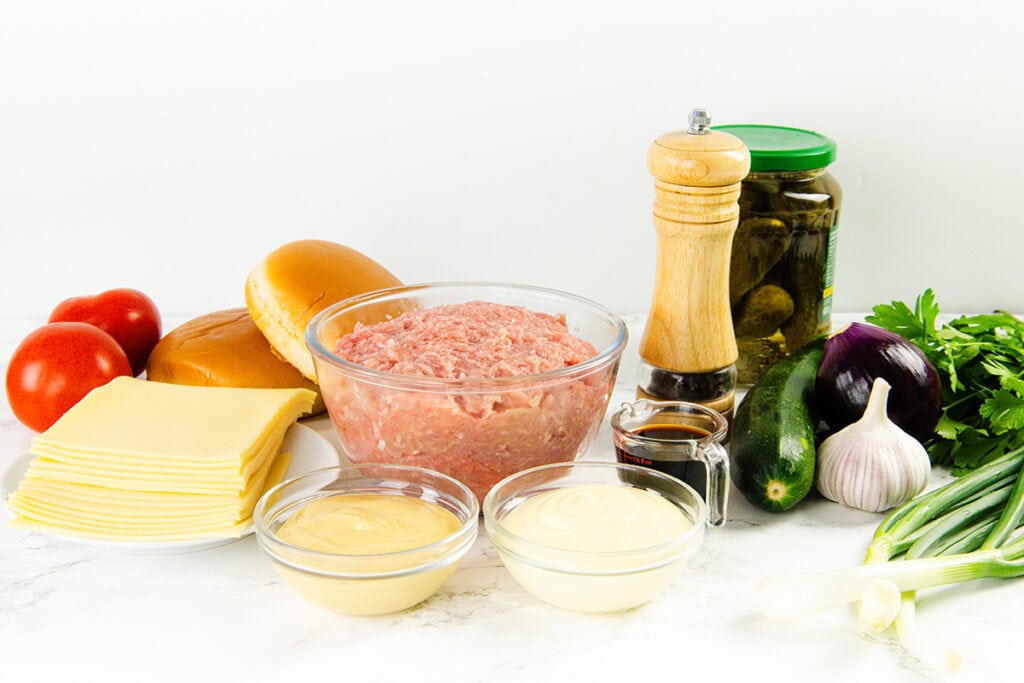 A variety of burger ingredients on a white surface, including ground meat for moist turkey burgers, cheese slices, tomatoes, burger buns, pickles, mustard, mayonnaise, a pepper mill, an onion, a cucumber, and fresh herbs.