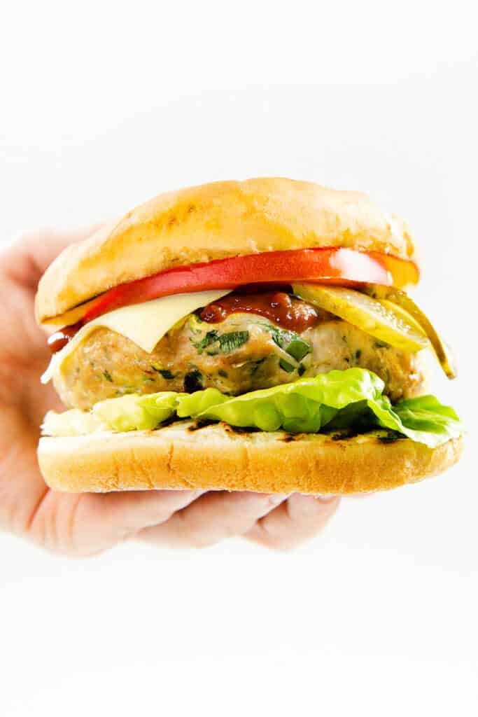 A hand holds a turkey burger with lettuce, cheese, tomato, pickles, and ketchup inside a soft bun, against a white background.