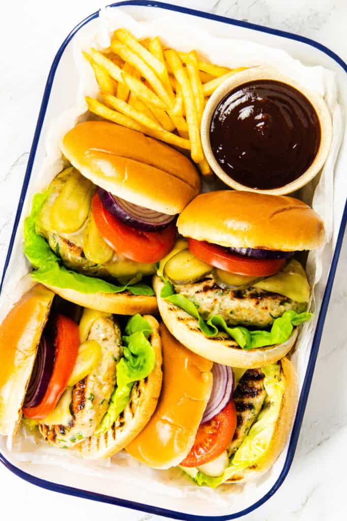 Four moist turkey burgers with lettuce, tomato, onion, and cheese in buns, served with a side of French fries and a small bowl of dark dipping sauce in a white tray.