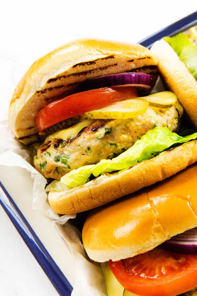 Close-up of a moist turkey burger in a soft bun with lettuce, tomato, red onion, pickles, and a grilled ground turkey patty, served on a tray with another similar burger partially visible.