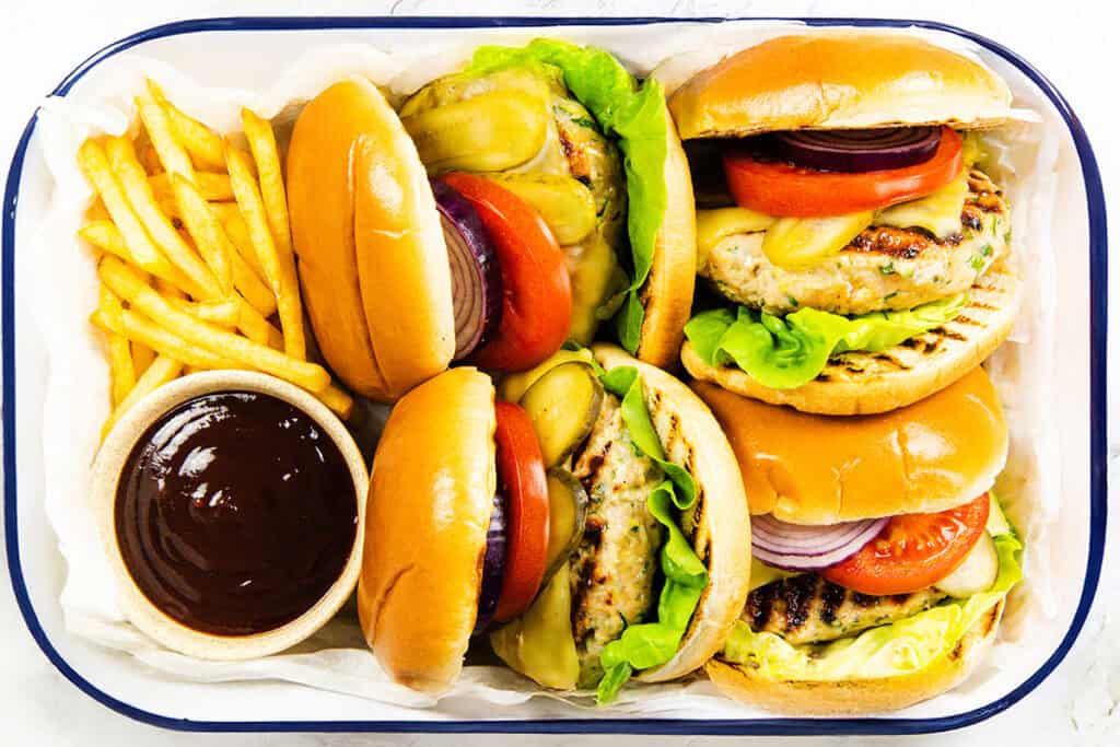 A tray with three moist turkey burgers on buns with lettuce, tomato, pickles, and onion, served alongside crispy French fries and a small bowl of dark dipping sauce.