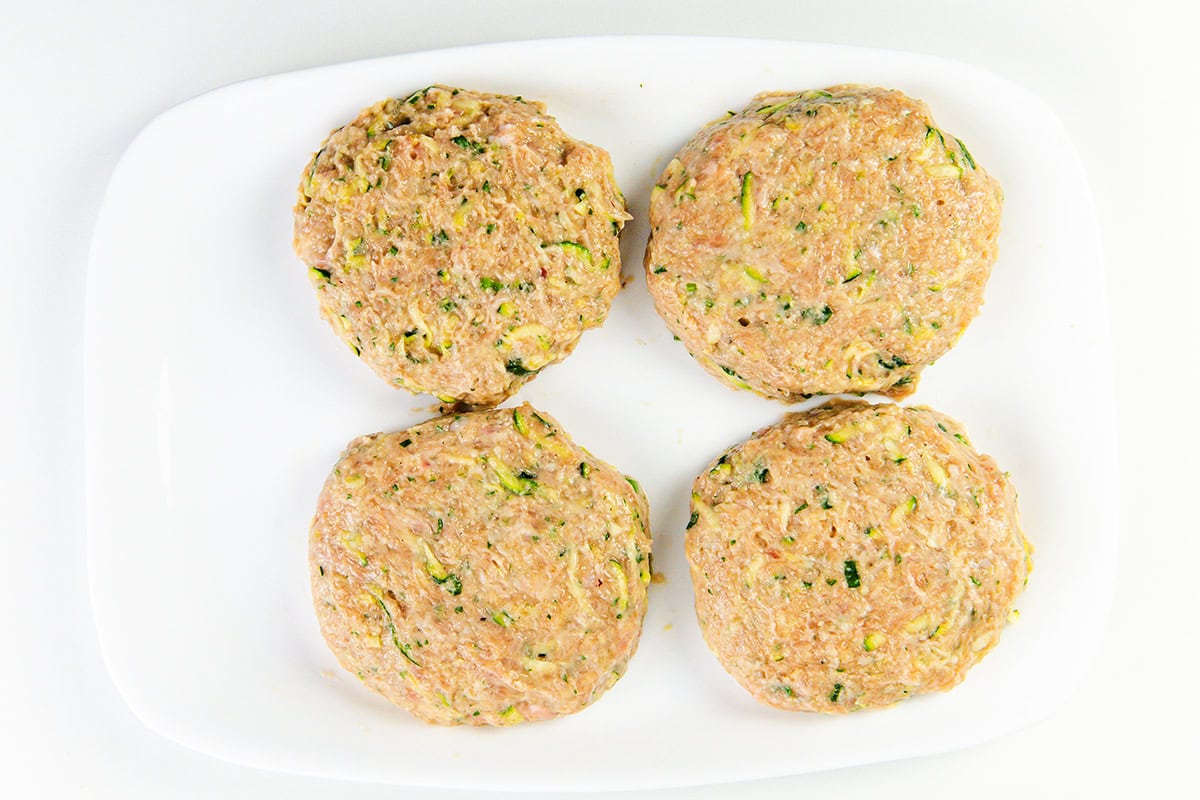 Four raw, round ground turkey burgers with visible green zucchini shreds are arranged on a white rectangular plate against a white background—perfect for your next moist turkey burger recipe.