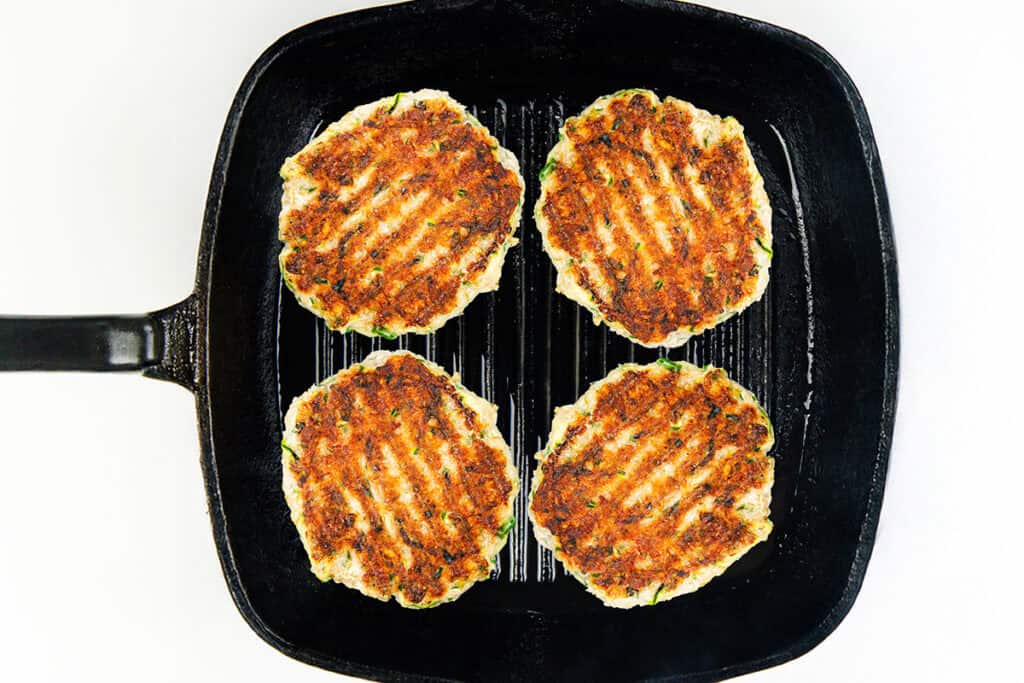 Four moist turkey burgers with perfect grill marks sizzle in a black grill pan, viewed from above, on a white background.
