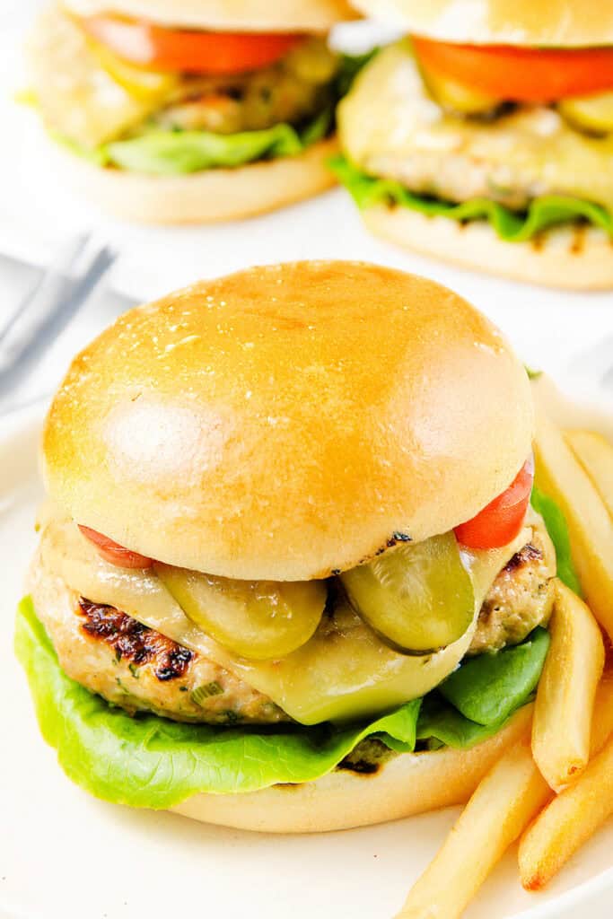 A close-up of a super moist ground turkey burger with lettuce, pickles, tomato, and cheese in a shiny bun, served with French fries. Two more burgers are blurred in the background, perfect inspiration for your next turkey burger recipe.