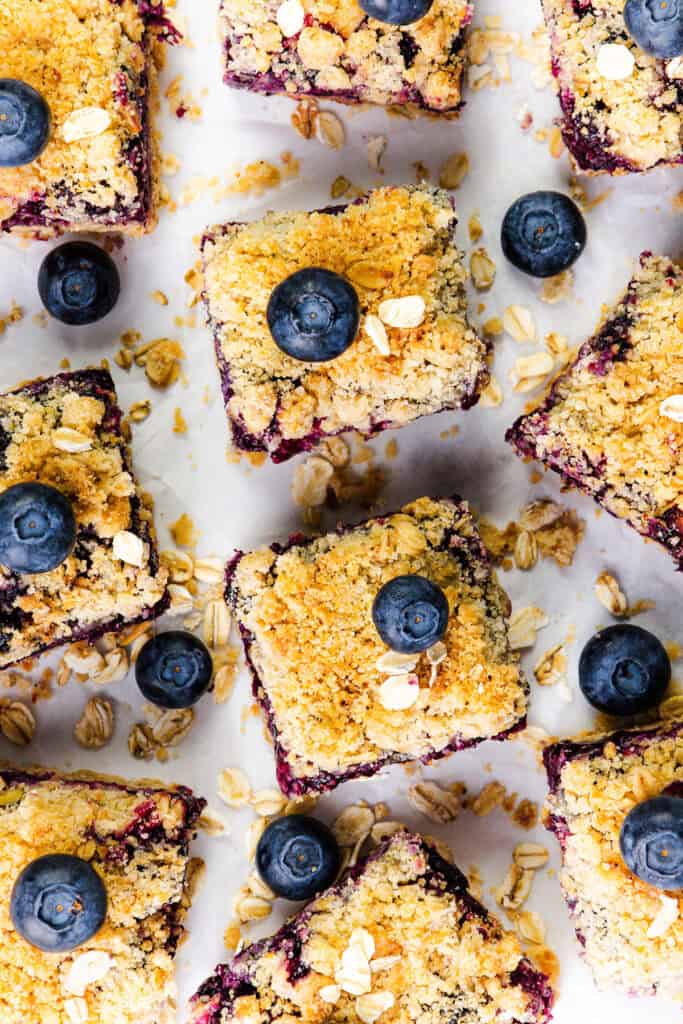 Top-down view of Blueberry Oatmeal Bars scattered on a white surface, topped with fresh blueberries and oats. The bars feature a golden crumb topping and a rich blueberry filling.