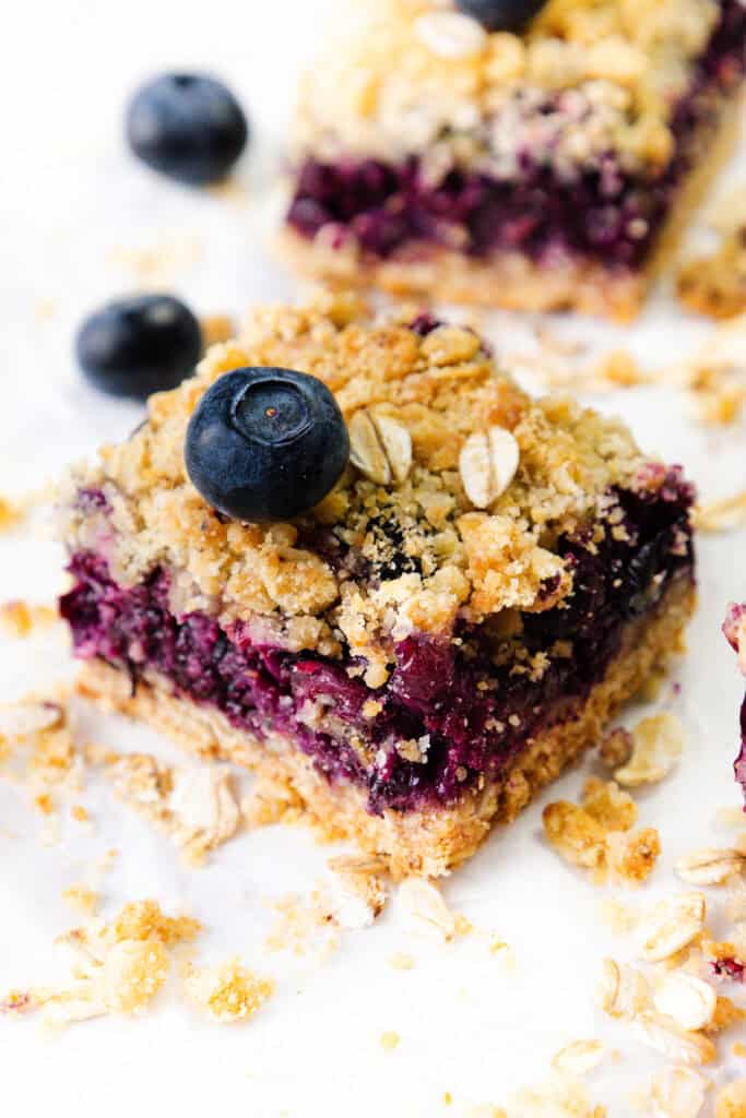 A blueberry crumb bar, reminiscent of classic Blueberry Bars, topped with a fresh blueberry and oat crumble, surrounded by scattered oats and crumbs on a white surface.