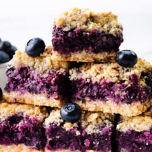 A stack of Blueberry Oatmeal Bars topped with fresh blueberries sits on a white surface, with a cup of blueberries in the background.