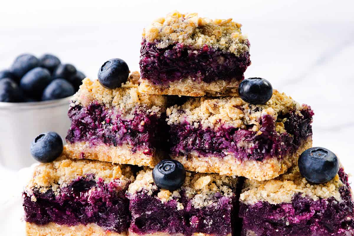A stack of Blueberry Oatmeal Bars topped with fresh blueberries sits on a white surface, with a cup of blueberries in the background.
