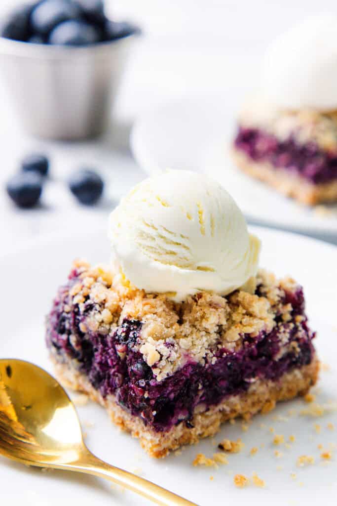 A square Blueberry Oatmeal Bar topped with a scoop of vanilla ice cream sits on a white plate next to a gold spoon, while a bowl of fresh blueberries is blurred in the background.