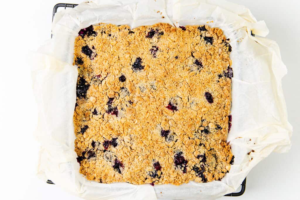 A square pan lined with parchment paper holds golden brown Blueberry Oatmeal Bars, featuring visible pieces of dark blueberries and a crumbly oat topping.