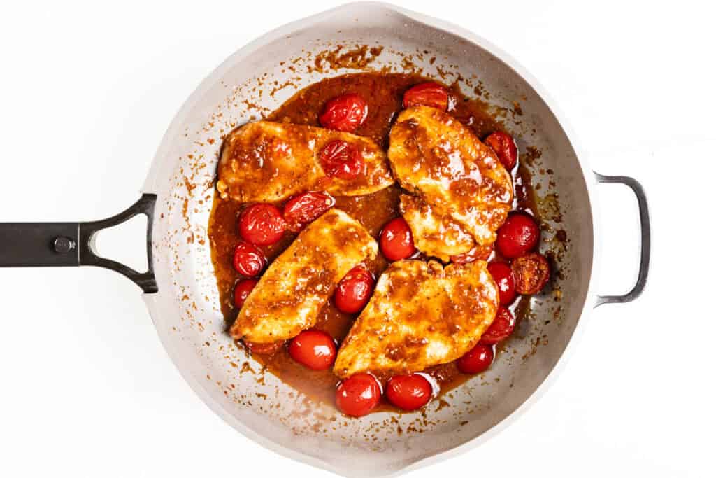 Four cooked Caprese Chicken breasts with a glossy sauce and cherry tomatoes in a white skillet, viewed from above on a white background.