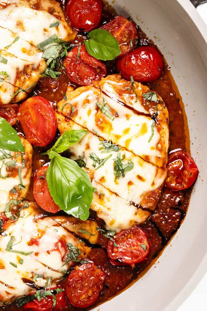 Caprese Chicken breasts topped with melted cheese, sliced cherry tomatoes, tomato sauce, and fresh basil leaves in a skillet, garnished with chopped herbs.