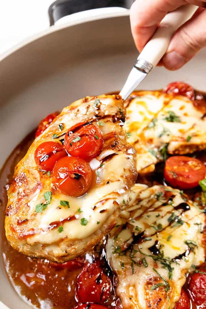 A hand holds a spatula lifting a piece of Caprese Chicken breast topped with melted cheese and roasted cherry tomatoes from a pan, with more chicken and tomatoes visible below.