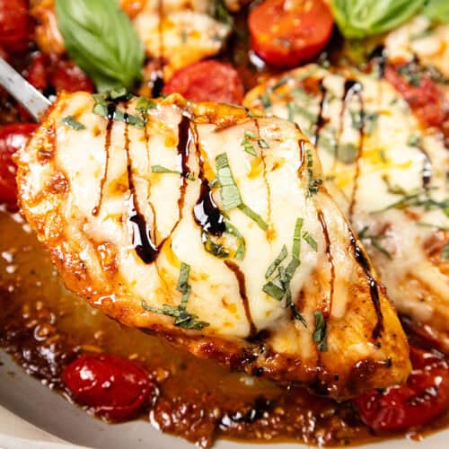 A close-up of Caprese Chicken: a cheesy baked chicken breast topped with fresh basil and a drizzle of balsamic glaze, served with roasted cherry tomatoes in a pan.